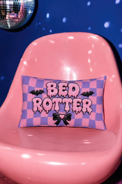 Pastel goth lumbar pillow, 20 by 12 inches, with a pink and lavender purple wavy checkerboard background. The words Bed Rotter appear in dripping pink bubble letters with a faux embroidery thread texture and black outlines. Two black bats with pink stitch details flank the text, and a black bow with a pink heart center sits below. Double sided flat print that mimics raised embroidery on linen feel polyester with a hidden zipper, insert included.
