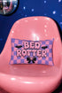 Pastel goth lumbar pillow, 20 by 12 inches, with a pink and lavender purple wavy checkerboard background. The words Bed Rotter appear in dripping pink bubble letters with a faux embroidery thread texture and black outlines. Two black bats with pink stitch details flank the text, and a black bow with a pink heart center sits below. Double sided flat print that mimics raised embroidery on linen feel polyester with a hidden zipper, insert included.