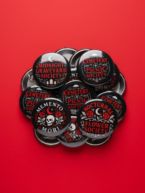 A collection of four glossy black circular pinback buttons featuring striking red and white gothic illustrations. One button shows "Midnight Graveyard Society" text with bats and tombstones. Another reads "Cemetery Picnic Society" with a coffin and umbrella icon. A third features a detailed white skull and red roses with "Memento Mori" text below. The final pin displays "Nocturnal Flower Society" surrounded by floral vines.