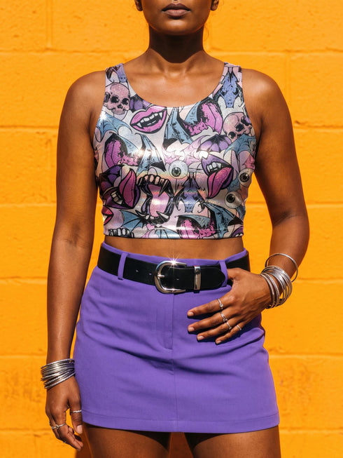 A sleeveless cropped tank top made from stretchy performance knit fabric. The all-over print features a dense, scattered pattern of pastel goth and creepy cute motifs over a light background. Graphics include pink and blue bats in flight, floating eyeballs with blue irises, pink skulls, and glossy vampire lips with sharp white fangs and tongues sticking out. The neckline is a high scoop and the armholes are curved. The fabric comes in either a matte finish or a sparkly silver glitter foil.