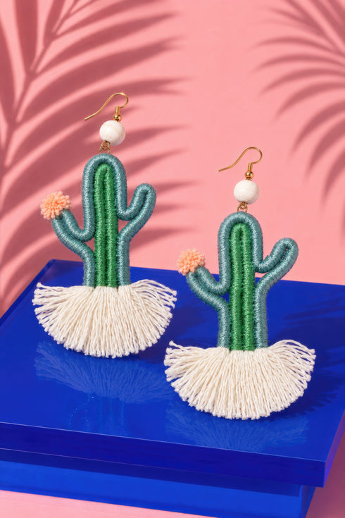a pair of earrings shaped like cacti, with green and white fringed bases and white beads hanging from the top. The earrings are displayed on a blue surface against a pink background.