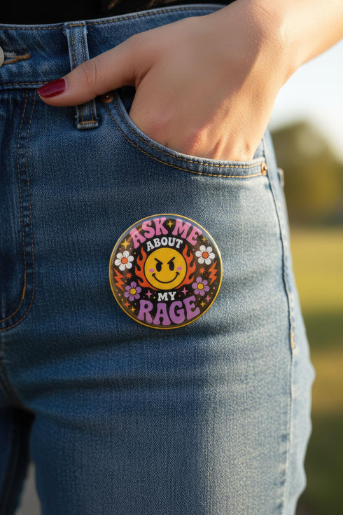 A round glossy metal pinback button featuring a flat 2D print design on a solid black background. In the center is an angry yellow smiley face surrounded by orange and red flames. Above the smiley face, pink text reads ASK ME and white text reads ABOUT. Below the face, white text reads MY and large purple text reads RAGE. The design is accented with two white daisies, two purple flowers, pink and orange four-point stars, and orange lightning bolts framing the outer edges.