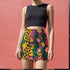 A woman wearing a black crop top and a colorful, patterned mini skirt stands against a pink wall.