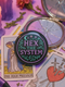 Glossy black pinback button with pastel embroidery-style design that says “Hex the System,” surrounded by vines, stars, and a crescent moon. Displayed against witchy pink and blue backdrops, street pole flyers, and protest props.