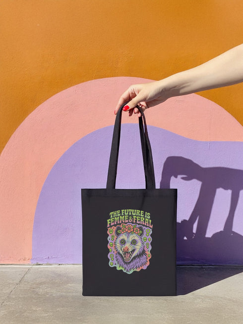 Black or natural cotton canvas tote bag with “The Future is Femme & Feral” design featuring a rainbow-colored snarling opossum with flowers and feminist symbols, printed on both sides with long matching canvas handles.
