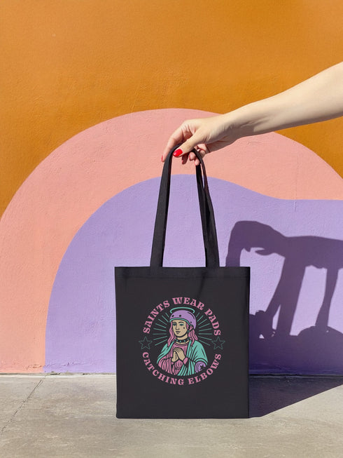 Black cotton canvas tote bag featuring a bold graphic of a pink-haired saint wearing a helmet, surrounded by the text “Saints Wear Pads Catching Elbows” in pastel pink and green gothic-style lettering. The tote has reinforced self-fabric handles and flat corners, ideal for daily use or carrying roller skating gear.