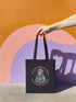 Black cotton canvas tote bag featuring a bold graphic of a pink-haired saint wearing a helmet, surrounded by the text “Saints Wear Pads Catching Elbows” in pastel pink and green gothic-style lettering. The tote has reinforced self-fabric handles and flat corners, ideal for daily use or carrying roller skating gear.