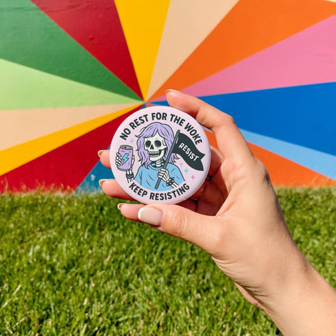 Honey Dazed Glossy round pink pinback button featuring a skeleton girl with purple hair holding a black flag that says “RESIST” and an energy drink can, with the phrases “No Rest for the Woke” and “Keep Resisting” in bold black letters.
