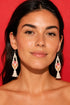 A woman with long dark hair wearing large, dangling earrings against a red background.