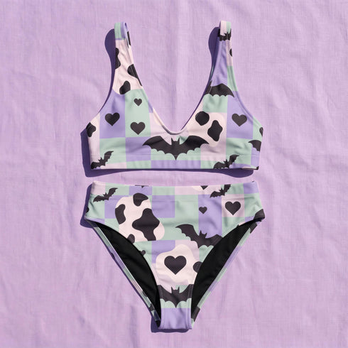 A two piece recycled high waisted swimsuit with a pastel goth checkered print in light lavender purple, pale mint green, and white squares with black cow print spots. The design features small black hearts and silhouetted bats with outspread wings printed throughout. The athletic style crop top has a deep scoop neck and matching thick straps, while the double layered bottoms have a supportive high waistband and cheeky cut with a solid black inner lining.