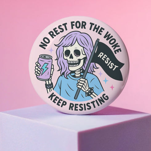 Honey Dazed Glossy round pink pinback button featuring a skeleton girl with purple hair holding a black flag that says “RESIST” and an energy drink can, with the phrases “No Rest for the Woke” and “Keep Resisting” in bold black letters.