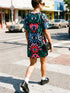A short-sleeve, above-the-knee t-shirt dress with a true black background. The front features a vibrant, vertically stacked symmetrical graphic design. From top to bottom, the design includes a pink heart containing a green and blue eye, a large blue sunburst with a central eye, a red daisy flower with an eye flanked by pink and black checkered arches, and a pink mushroom cap with an eye. Swirling emerald green foliage, pink stars, and purple stars fill the negative space around the motifs.