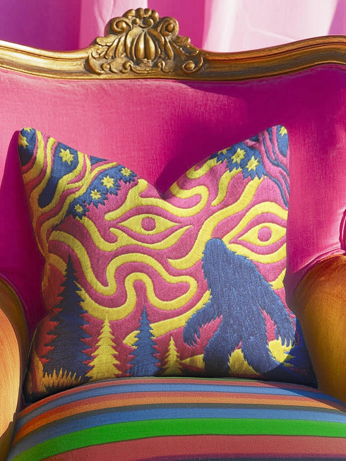 A square decorative throw pillow cover featuring a trippy retro Bigfoot scene. A large navy blue silhouette of Sasquatch walks through a forest of navy and yellow pine trees. The background is a psychedelic swirling sky in vibrant pink and yellow, featuring a large stylized eye and a cluster of small yellow stars. The flat two dimensional print visually mimics the textured look of heavy needlepoint embroidery. The pillow features a sleek hidden zipper on the bottom. No insert is included.