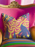 A square decorative throw pillow cover featuring a trippy retro Bigfoot scene. A large navy blue silhouette of Sasquatch walks through a forest of navy and yellow pine trees. The background is a psychedelic swirling sky in vibrant pink and yellow, featuring a large stylized eye and a cluster of small yellow stars. The flat two dimensional print visually mimics the textured look of heavy needlepoint embroidery. The pillow features a sleek hidden zipper on the bottom. No insert is included.