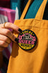 Round pinback button badge with Local Queer Menace text in bold yellow and pink retro lettering on a black background. A cartoon raccoon with a spiked collar flips the middle finger with a painted pink nail, framed by a hot pink sunburst, lightning bolts, and star accents. Glossy laminate finish over durable metal tinplate with a secure safety pin backing. Funny queer pride pin available in 2.25 inch and 3 inch sizes for jackets, totes, lanyards, and backpacks.