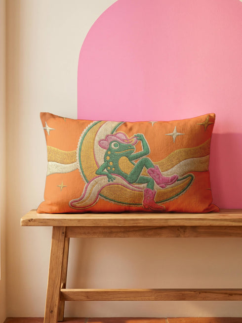 Rectangular 20x12 inch lumbar pillow cover in bright orange featuring printed faux embroidery of a green frog wearing a pink cowgirl hat and pink cowboy boots, lounging on a golden yellow crescent moon shaped like a peeled banana, surrounded by cream and gold sparkle stars and retro wavy stripes. Whimsical cowboy frog design printed on both sides with a hidden zipper, crafted from soft linen look polyester in a funky 70s retro style.