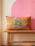 Rectangular 20x12 inch lumbar pillow cover in bright orange featuring printed faux embroidery of a green frog wearing a pink cowgirl hat and pink cowboy boots, lounging on a golden yellow crescent moon shaped like a peeled banana, surrounded by cream and gold sparkle stars and retro wavy stripes. Whimsical cowboy frog design printed on both sides with a hidden zipper, crafted from soft linen look polyester in a funky 70s retro style.