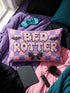 Pastel goth lumbar pillow, 20 by 12 inches, with a pink and lavender purple wavy checkerboard background. The words Bed Rotter appear in dripping pink bubble letters with a faux embroidery thread texture and black outlines. Two black bats with pink stitch details flank the text, and a black bow with a pink heart center sits below. Double sided flat print that mimics raised embroidery on linen feel polyester with a hidden zipper, insert included.