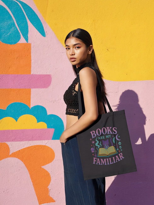Black cotton tote bag with pastel goth design featuring “Books Are My Familiar” in soft pink, lavender, and mint tones. Design includes an open book with smoke, a silhouetted cat, moons, keys, stars, and magical icons. Shown in front of colorful mural walls and worn by models in alternative fashion. Double-sided print, perfect for witchy readers, soft goths, and literary magic lovers.