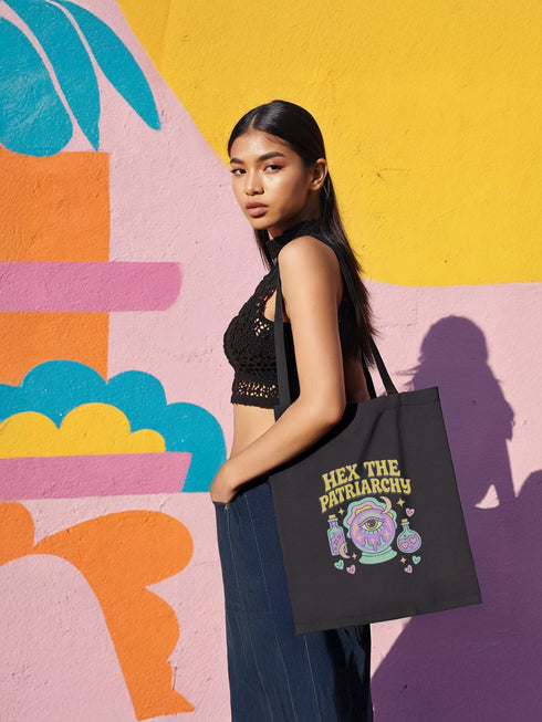 Canvas cotton tote bag with bold “Hex the Patriarchy” design printed on the front, featuring a dripping purple eye in a crystal ball surrounded by pastel potion bottles and hearts. Bag is available in black or natural cotton with long canvas handles.