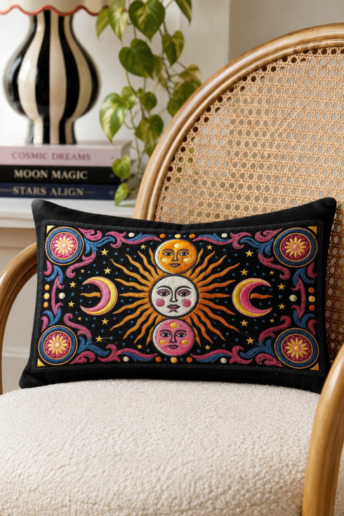 Rectangular 20x12 inch lumbar pillow cover in black featuring printed faux crewel embroidery of a large orange sunburst with a cream face, flanked by pink and yellow crescent moons, a golden moon face above and a pink moon face below, gold stars, blue dots, magenta and blue baroque scroll flourishes, and four corner medallions with golden flowers. Whimsigoth celestial design printed on both sides with a hidden zipper, crafted from soft linen look polyester.