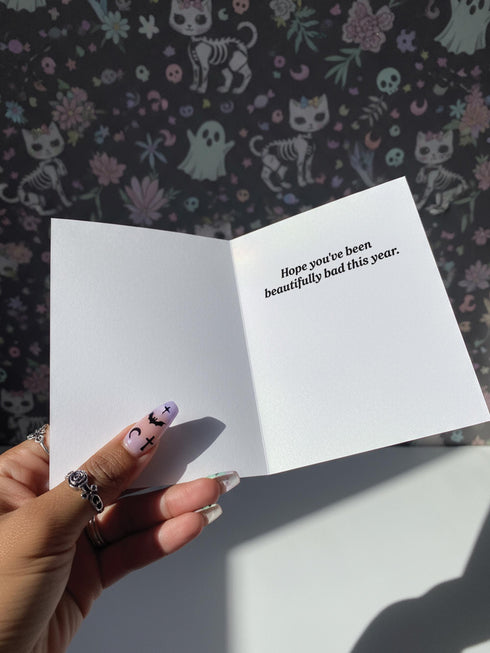 A 5x7 inch Krampus Christmas card shown in two paper finishes. The front features a stylized illustration of the folklore demon Krampus with green skin, black horns, and a pink Santa hat. He carries a large pink sack filled with three skulls over his shoulder. Below him, the words "KRAMPUS IS COMING" are printed in bold pink text. The inside of the card is blank on the left and has the printed text "Hope you've been beautifully bad this year." on the right.