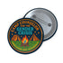 Round glossy pinback button measuring 2.25 inches featuring a playful camp-themed design with the phrase “Camp Counselor for the Gender Crisis.” The artwork includes two orange tents, a rainbow campfire, stars, and green grass. Bottom text reads “Cabin Queer, Unit Psychedelic.” Made from durable tinplate with a safety pin back and coated in a UV and scratch-resistant mylar finish. Bold and colorful, perfect for jackets, bags, or pride outfits.