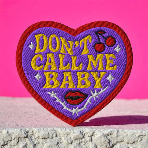 A heart-shaped embroidered patch with a thick red border and a vibrant purple background. In the center, the phrase "DON’T CALL ME BABY" is stitched in a bold, golden-yellow block font. The patch is decorated with small white sparkles, a pair of red cherries with a black musical note, a pair of red lips, and a grey thorny vine design below the text. The overall design has a rebellious, retro, punk aesthetic with a handmade, textured look.
