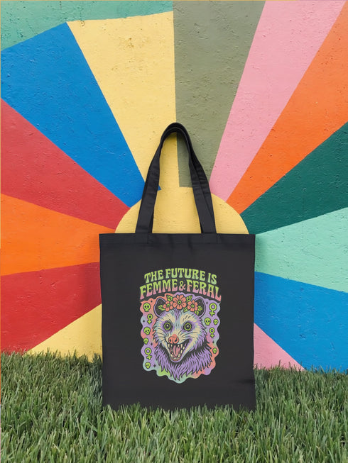 Black or natural cotton canvas tote bag with “The Future is Femme & Feral” design featuring a rainbow-colored snarling opossum with flowers and feminist symbols, printed on both sides with long matching canvas handles.