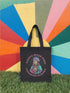 Black cotton canvas tote bag featuring a bold graphic of a pink-haired saint wearing a helmet, surrounded by the text “Saints Wear Pads Catching Elbows” in pastel pink and green gothic-style lettering. The tote has reinforced self-fabric handles and flat corners, ideal for daily use or carrying roller skating gear.