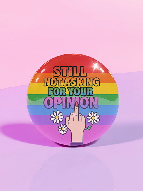 Round glossy pinback button measuring 2.25 inches with a bold rainbow pride flag background. Centered text reads “Still Not Asking For Your Opinion” in black, yellow, blue, and purple letters. Below the text is an illustrated hand with painted red nails giving the middle finger, surrounded by white daisies. The design is printed on tinplate with a scratch- and UV-resistant mylar coating. Button features a strong safety pin backing, perfect for bags, jackets, or pride outfits.