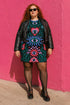 A short-sleeve, above-the-knee t-shirt dress with a true black background. The front features a vibrant, vertically stacked symmetrical graphic design. From top to bottom, the design includes a pink heart containing a green and blue eye, a large blue sunburst with a central eye, a red daisy flower with an eye flanked by pink and black checkered arches, and a pink mushroom cap with an eye. Swirling emerald green foliage, pink stars, and purple stars fill the negative space around the motifs.