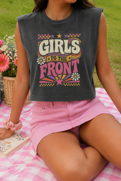A women's garment-dyed muscle tee in faded charcoal grey, featuring a boxy, slightly cropped fit with structured shoulders and a wide ribbed collar. The center chest displays a retro graphic reading GIRLS TO THE FRONT in cream and magenta bubbly typography. The text is surrounded by vintage motifs including white daisies, yellow and purple stars, wavy ribbon banners, pink and yellow checkerboard borders, and a bottom radial sunburst in orange and magenta. Made of one hundred percent cotton.