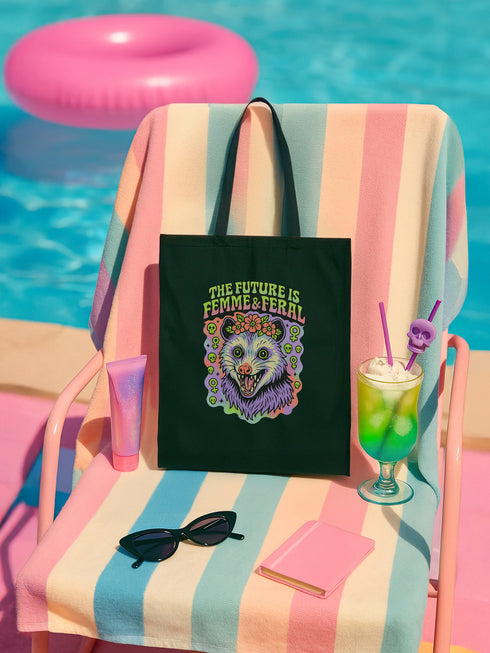 Black or natural cotton canvas tote bag with “The Future is Femme & Feral” design featuring a rainbow-colored snarling opossum with flowers and feminist symbols, printed on both sides with long matching canvas handles.