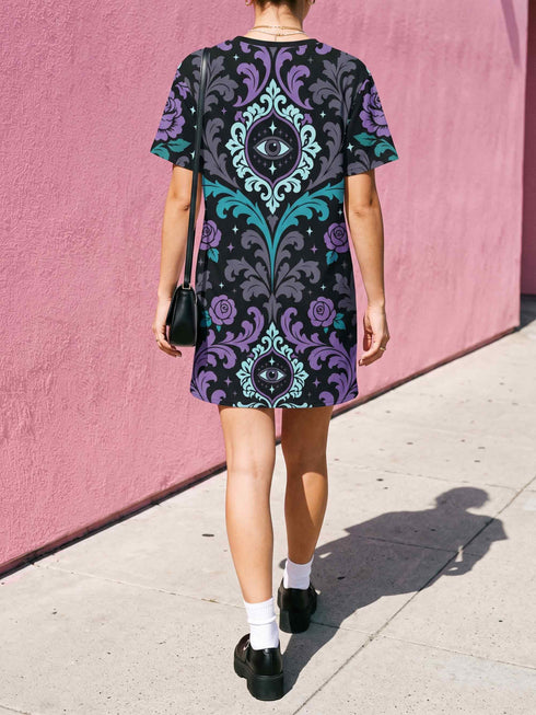 Cotton t-shirt dressA short-sleeve structured t-shirt dress with a black background. The dress features a bold, vertically stacked repeating graphic print. The design includes intricate damask patterns in teal and grey, purple roses with teal leaves, and a central evil eye motif set inside an ornate frame with small four-point stars. The heavyweight fabric holds a clean, straight cut above the knee with zero drape, maintaining a sculptural, slightly boxy silhouette.