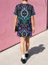 Cotton t-shirt dressA short-sleeve structured t-shirt dress with a black background. The dress features a bold, vertically stacked repeating graphic print. The design includes intricate damask patterns in teal and grey, purple roses with teal leaves, and a central evil eye motif set inside an ornate frame with small four-point stars. The heavyweight fabric holds a clean, straight cut above the knee with zero drape, maintaining a sculptural, slightly boxy silhouette.