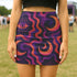 High-waisted mini skirt featuring a surreal wavy pattern with all-seeing eye and crescent moon motifs in magenta, plum, and burnt orange. Available in metallic or performance knit finishes. Sizes XS to XL.