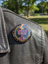 A round metal pinback button badge featuring a black background with the words QUEER MAGIC IS REAL in capital pink retro serif typography. A bright rainbow colored lightning bolt strikes from the top center, surrounded by concentric rainbow rings in red, orange, yellow, green, blue, and purple. The design is accented with small pink four-point stars and red crescent moons. The artwork features a realistic 2-D faux embroidery texture. The back has a silver metal safety pin clasp fastening.