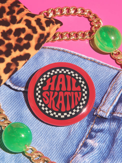 A round red pinback button with a glossy, scratch-resistant mylar face. The design features the phrase "HAIL SKATIN'" in a wavy, 70s psychedelic font in the center. This text is surrounded by a bold, black and white checkerboard pattern border, giving it a punk rock aesthetic. This piece of vintage-inspired flair is made of lightweight metal with a durable safety pin backing on the reverse. The button is a perfect for a denim jacket, backpack, or bag for fans of roller derby and skateboarding.