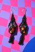 A pair of black earrings with a potted succulent plant hanging from the bottom, set against a vibrant background of pink and purple squares.