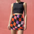 Honey Dazed mini pencil skirt featuring a bold wavy checkerboard pattern in black, orange, purple, and pink. Made from soft eco-polyester with a curve-hugging stretch fit and a flattering high-waist silhouette. The print has a slightly glossy finish, giving it a sleek and modern look. Designed for festival fits, streetwear moments, and retro rave energy. A vivid, sculptural mini skirt that pairs easily with crop tops and confident energy.