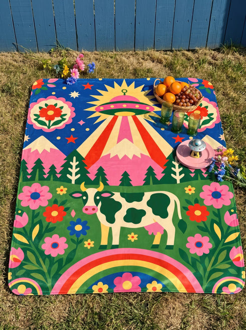 A rectangular velveteen minky blanket with a bright, maximalist design. The top half features a vibrant blue sky with a pink, green, and yellow UFO beaming rays down over hot pink snow-capped mountains. The bottom half shows a grassy green field with a white cow covered in dark spots, surrounded by large, colorful red, pink, and blue retro flowers, pine trees, and a rainbow arch at the very bottom. The reverse side is a fluffy off-white sherpa.