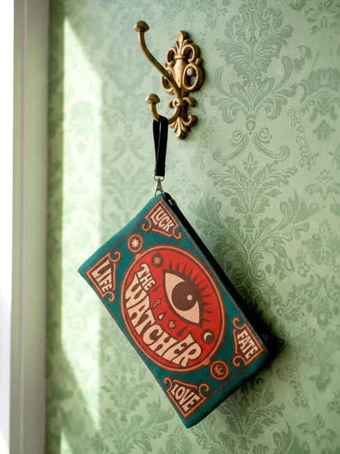 A rectangular 9x6 inch makeup bag and zipper pouch featuring a textured linen-like polyester fabric. The bag showcases a bold, maximalist print with vivid colors typical of retro 70s and pastel goth aesthetics, centering on a large mystic eye design labeled "The Watcher." It features a sturdy black zipper across the top and includes a removable black nylon wristlet strap.Perfect for use as a pencil case, cosmetic organizer, or festival wristlet.