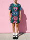 A short-sleeve, above-the-knee t-shirt dress with a true black background. The front features a vibrant, vertically stacked symmetrical graphic design. From top to bottom, the design includes a pink heart containing a green and blue eye, a large blue sunburst with a central eye, a red daisy flower with an eye flanked by pink and black checkered arches, and a pink mushroom cap with an eye. Swirling emerald green foliage, pink stars, and purple stars fill the negative space around the motifs.