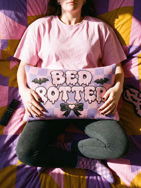 Pastel goth lumbar pillow, 20 by 12 inches, with a pink and lavender purple wavy checkerboard background. The words Bed Rotter appear in dripping pink bubble letters with a faux embroidery thread texture and black outlines. Two black bats with pink stitch details flank the text, and a black bow with a pink heart center sits below. Double sided flat print that mimics raised embroidery on linen feel polyester with a hidden zipper, insert included.