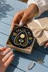 A close-up photograph of a glossy ceramic coaster featuring a Planchette occult design on a deep black background. The symmetrical graphic displays a central blue All-Seeing Eye with gold sunburst lines, topped by a white circle, and flanked by gold flowers with blue stems. At the top, the text "YES" is printed in gold on the left and "NO" on the right. Gold stars and crescent moons are scattered around the perimeter. The flat, round coaster has a highly visible high-gloss glaze.