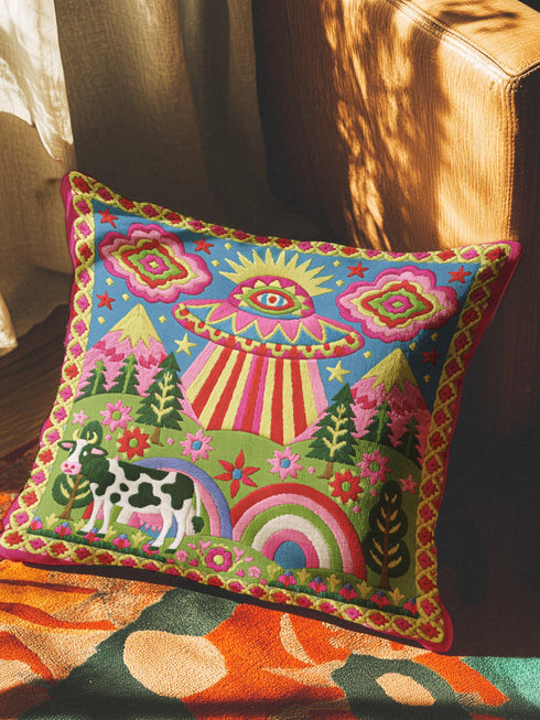 Description of a square throw pillow with a vibrant maximalist alien design. A central UFO with a large all-seeing eye hovers above a landscape with two rainbows, pink mountains, and green pine trees. A black and white spotted cow stands in the foreground. The design is a high-contrast palette of sky blue, magenta, hot pink, and meadow green. The pillow has a faux-embroidery 2D print finish on linen-feel fabric, framed by a yellow and pink scalloped floral border.