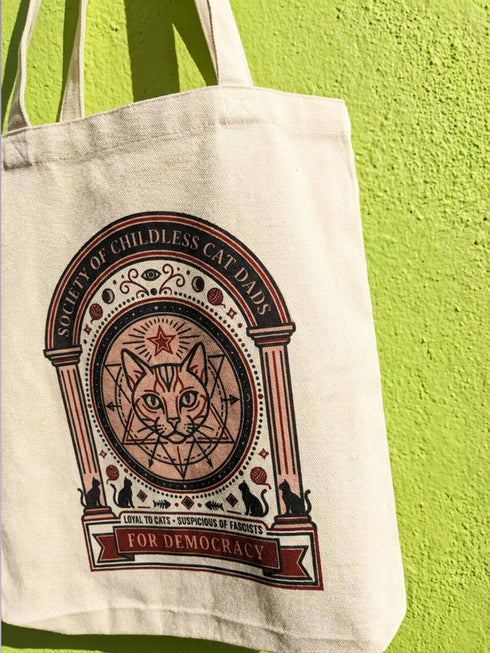 A beige canvas tote bag featuring a vintage-style circular crest design in rust orange and black. The text reads "Society of Childless Cat Dads" in an arch over a central illustration of a tabby cat face inside a geometric star and hexagon symbol. Below the cat, the text reads "Loyal to Cats, Suspicious of Fascists." A banner at the bottom reads "For Democracy." The design features occult and whimsigoth elements like crescent moons, columns, and fish bones.