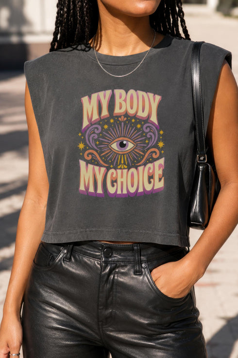 A garment dyed charcoal grey women's muscle tee featuring a slightly cropped boxy fit with structured shoulders and a wide rib collar. The front displays a retro 70s psychedelic graphic printed in purple, orange, and yellow. The text reads MY BODY MY CHOICE surrounding a central eye motif with a radial sunburst, framed by symmetrical mystical swirls and four point stars. The vintage style cotton shirt offers a faded aesthetic, presented on its own without background lifestyle elements.