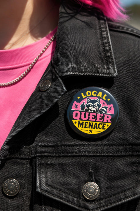 Round pinback button badge with Local Queer Menace text in bold yellow and pink retro lettering on a black background. A cartoon raccoon with a spiked collar flips the middle finger with a painted pink nail, framed by a hot pink sunburst, lightning bolts, and star accents. Glossy laminate finish over durable metal tinplate with a secure safety pin backing. Funny queer pride pin available in 2.25 inch and 3 inch sizes for jackets, totes, lanyards, and backpacks.
